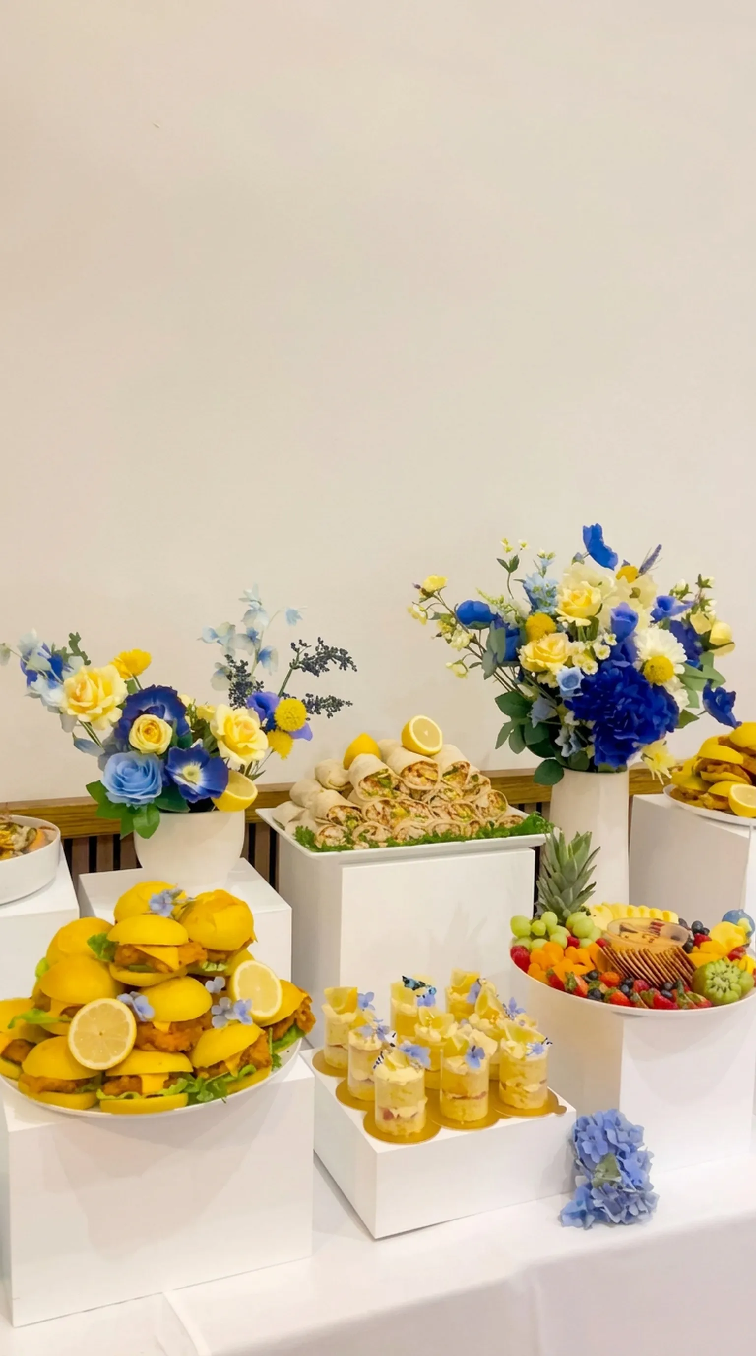 A blue and yellow corporate catering table with floral arrangements, savoury bites and cake jars